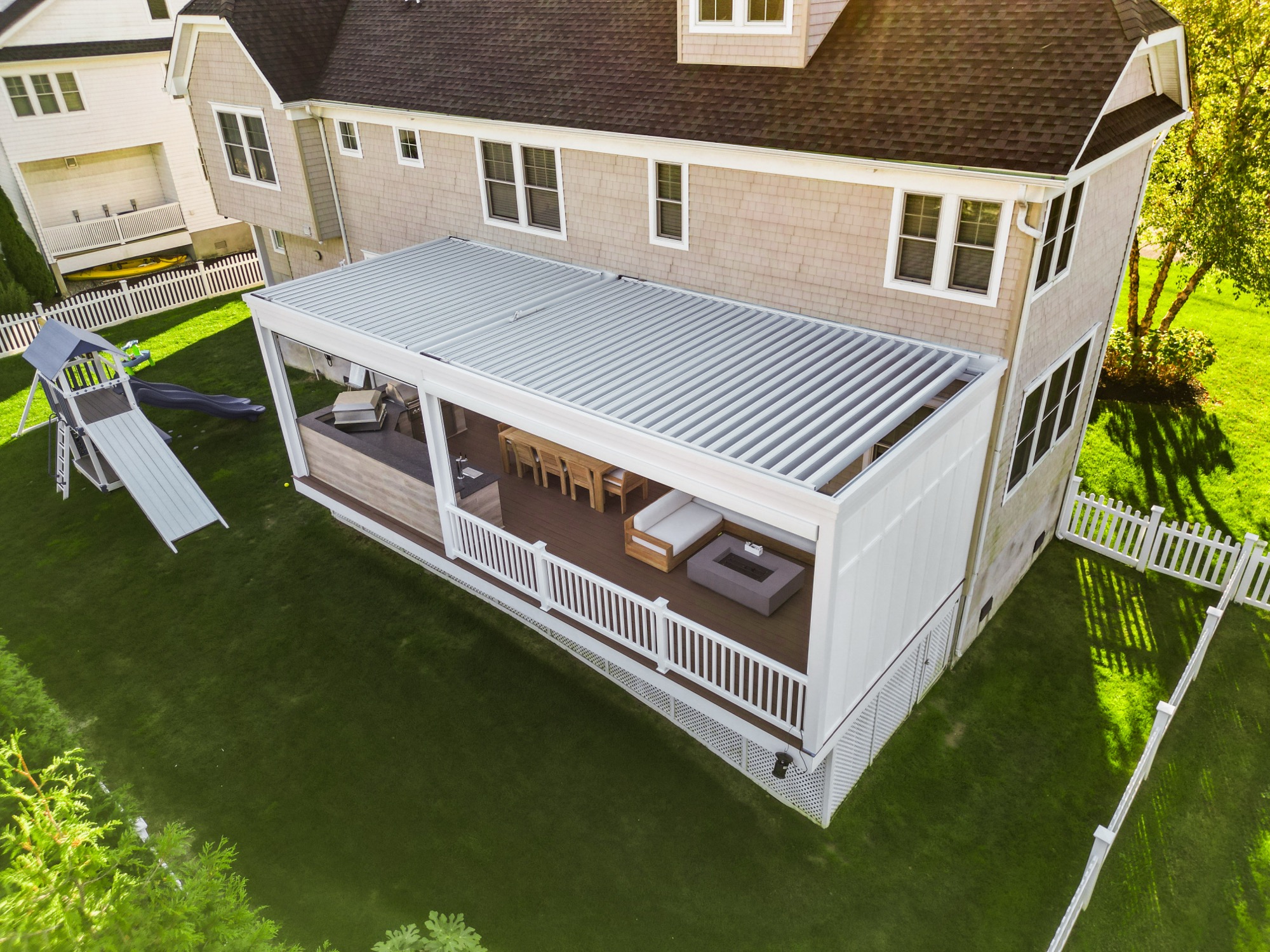 Aerial photo of a shingle-style house with a louvered roof over a deck with dining table, sofa, and outdoor kitchen