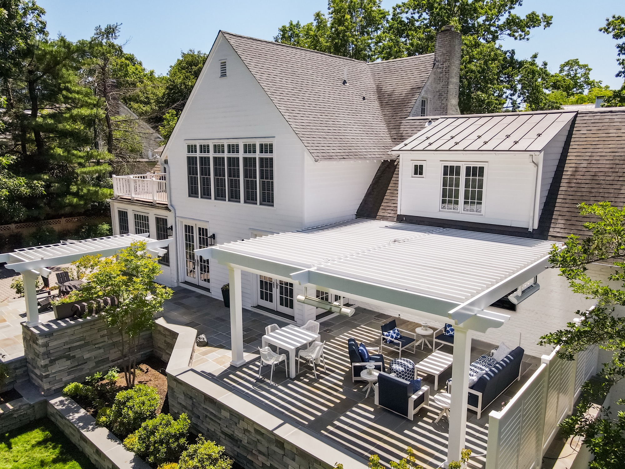 Large white shingle-style home with a louvered pergola over a stone patio with lounge furniture