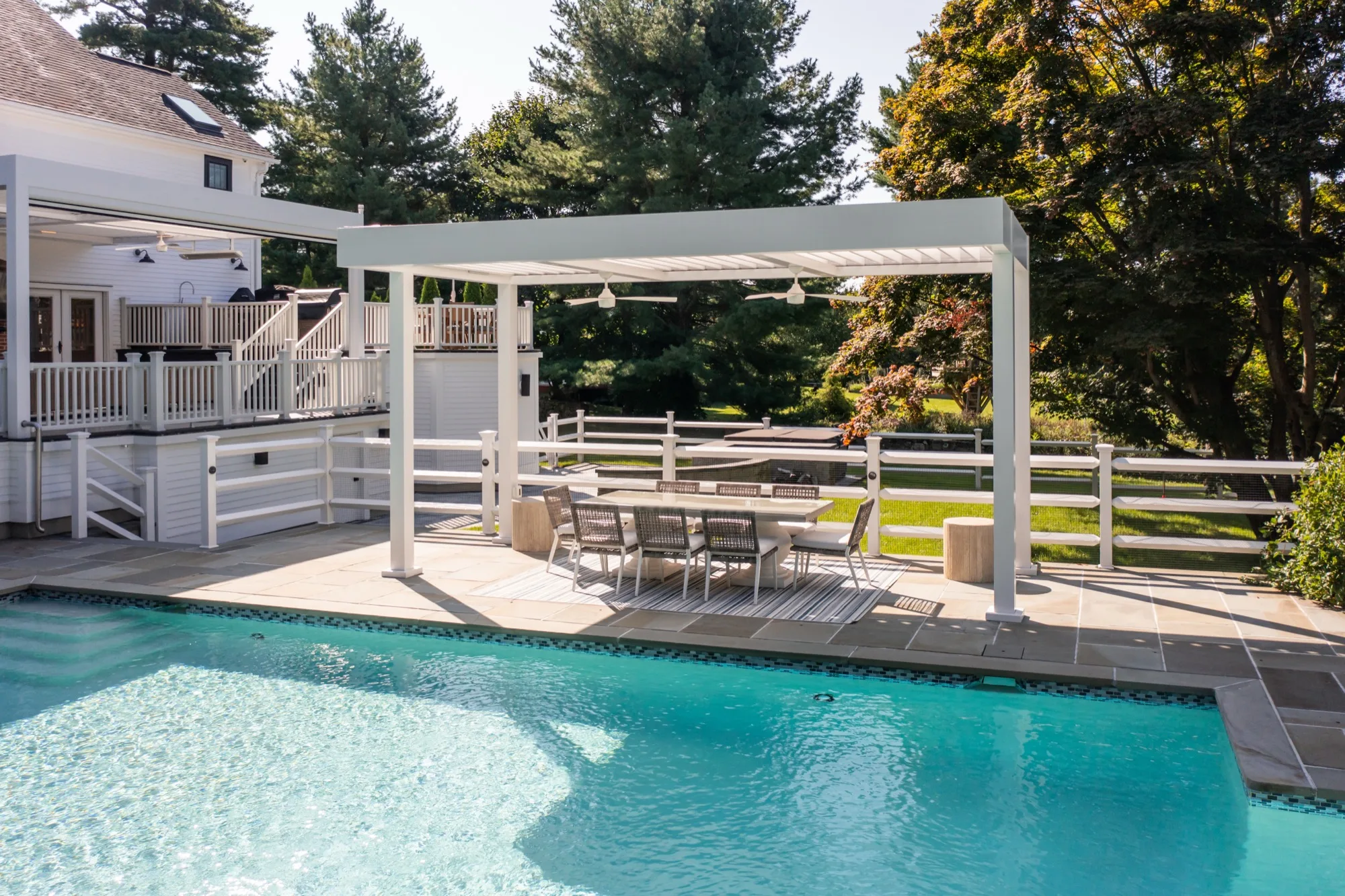 Aerial photo of a pool with a louvered pergola shading a dining set on the adjoining patio, autumn foliage visible