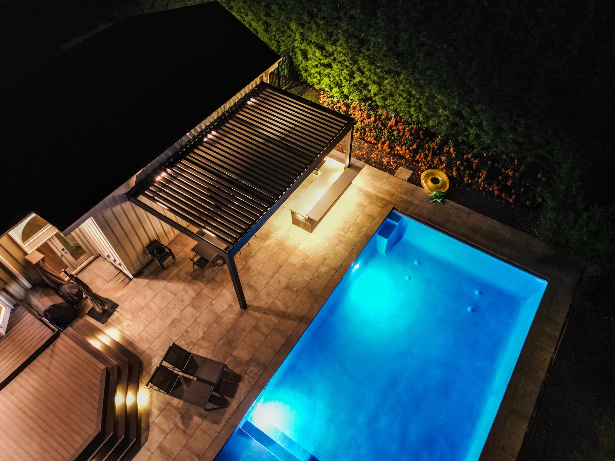 Nighttime aerial photo of an illuminated blue swimming pool beside a lit louvered pergola over a stone patio.