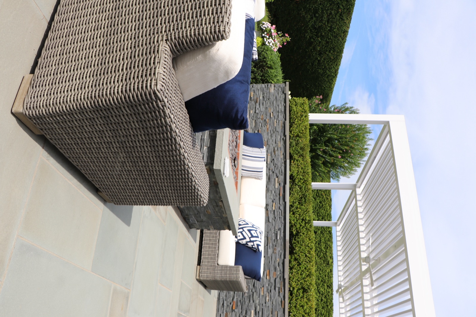 Wicker lounge seating with deep cream cushions and navy accent pillows beside the stone fire-pit table, the white double-bay Azenco R-Blade louvered roof and hedge wall behind — Southampton, NY