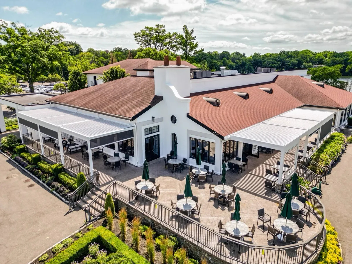 Aerial photo of a country club clubhouse with a louvered-pergola-covered patio, dining tables, and green umbrellas