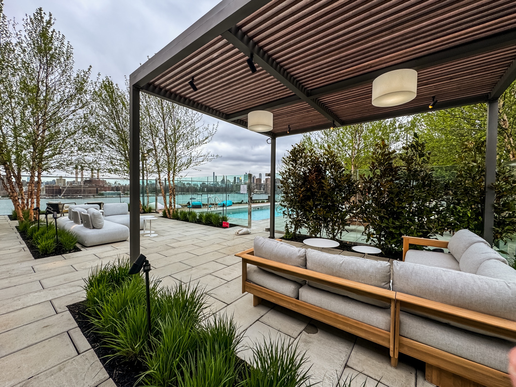 Rooftop lounge with a wood-slat pergola, white sofas, a pool, and a city skyline across the water