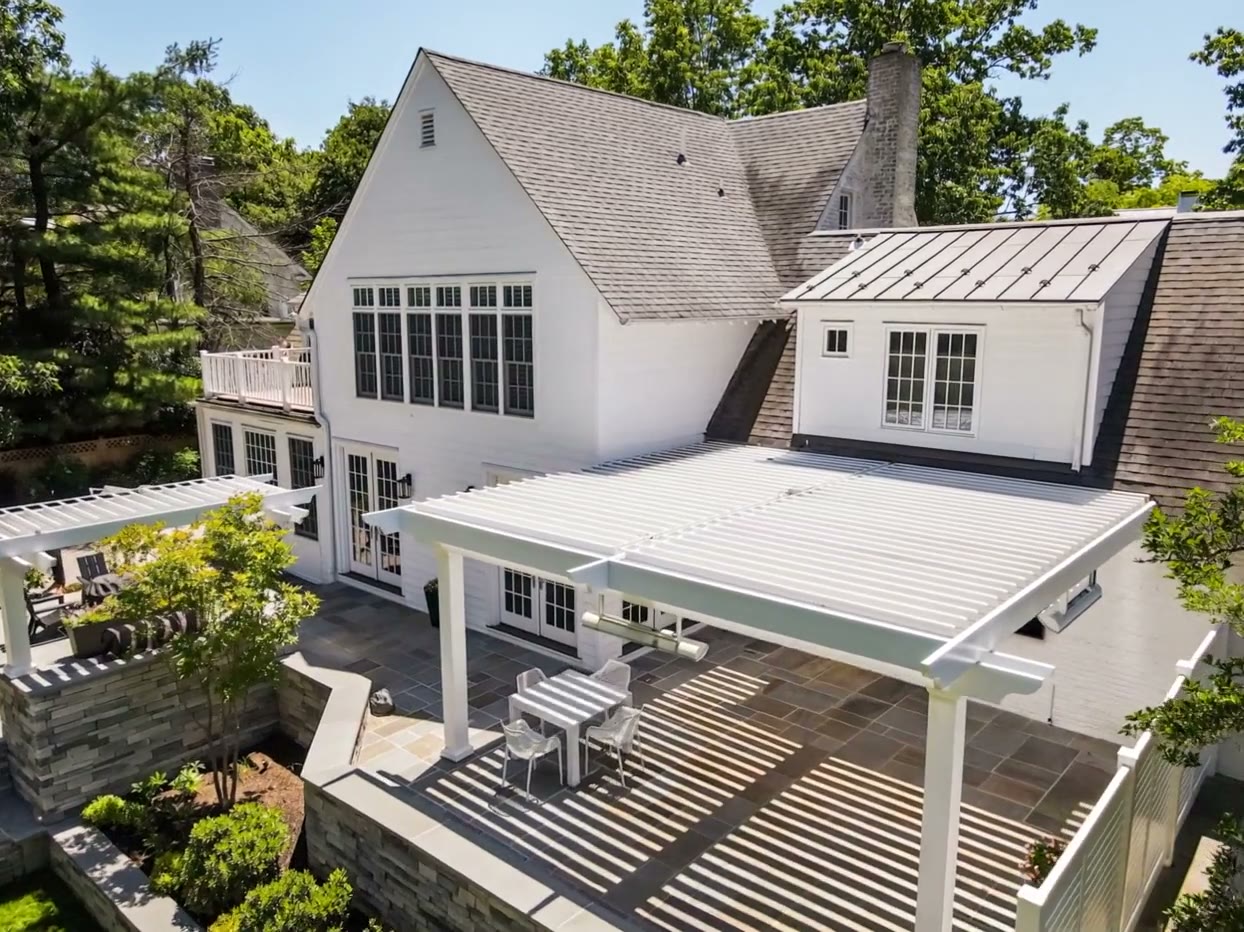 Aerial photo of a large white house with a louvered pergola over a stone patio holding a table and chairs
