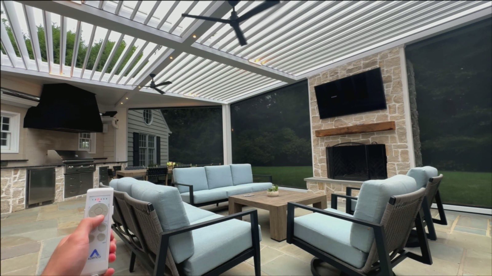 Covered patio with dark motorized screens lowered, stone fireplace, TV, and a hand holding an Azenco remote