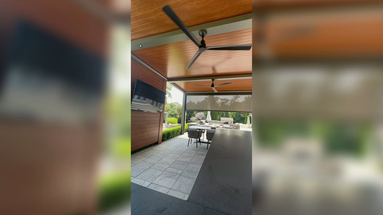 A wood-toned pergola ceiling with a fan and a partially lowered motorized screen over a stone patio