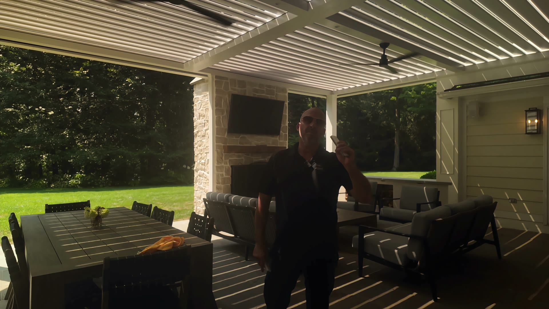 Interior view under a white louvered roof with an outdoor fireplace, TV, and lounge seating, showing the trimmed beam line where a shade can be concealed