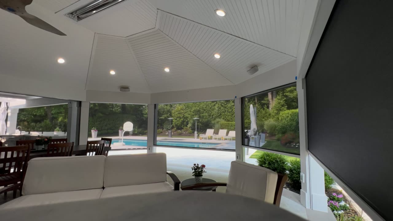 Photo of a screened pergola/porch interior with a pool visible outside, one motorized screen partially lowered, daytime