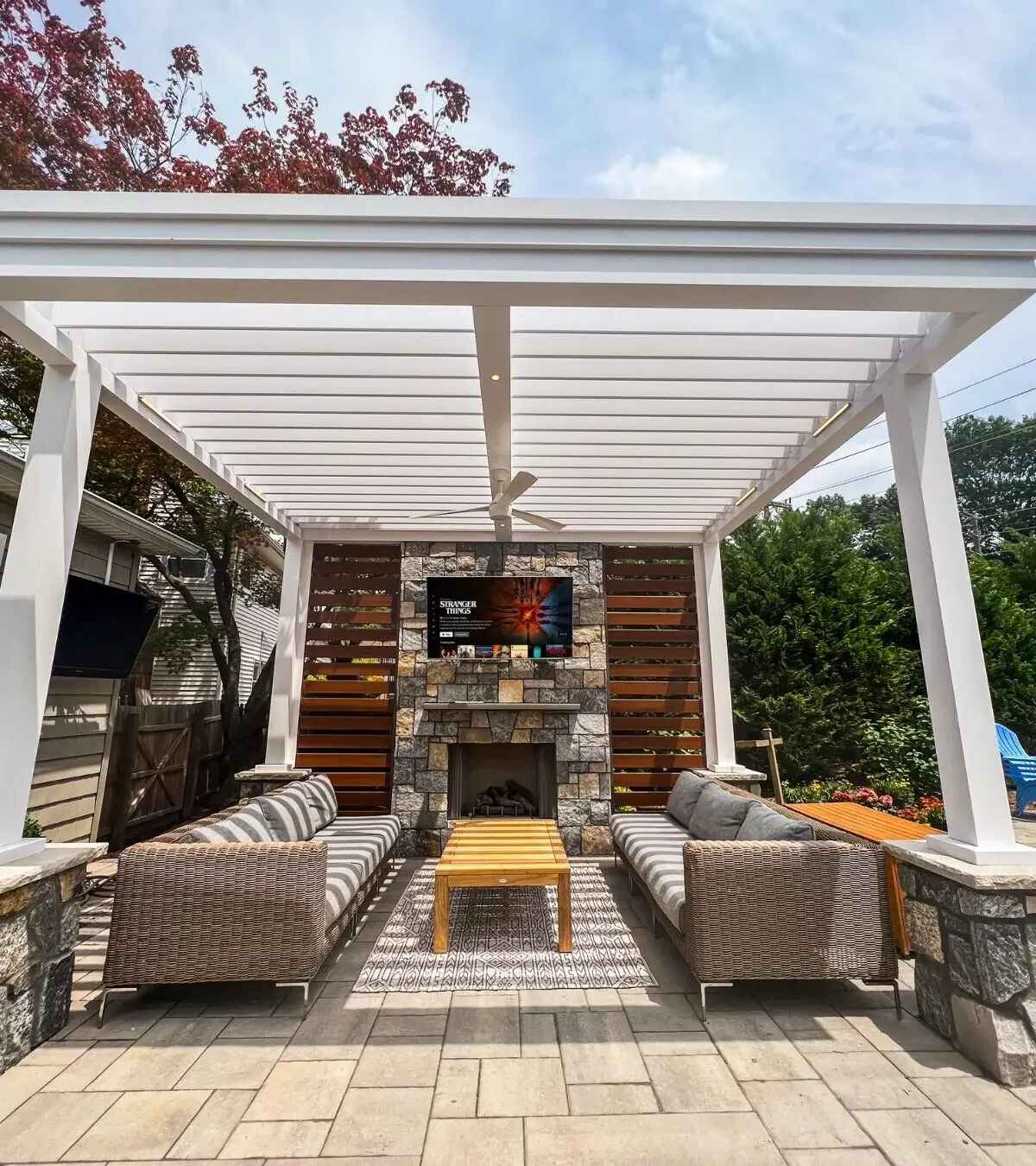 Photo of a white pergola-covered patio with wicker sofas, wood coffee table, and stone fireplace with mounted TV, autumn daytime.