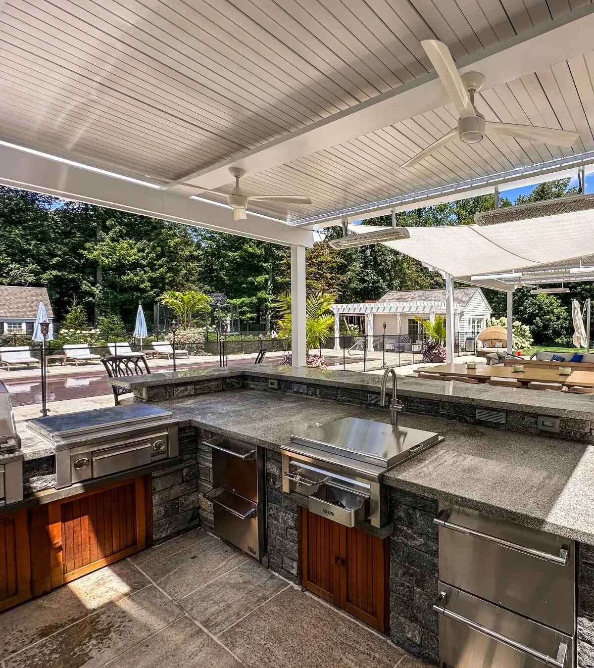 Outdoor kitchen counter with built-in grill, refrigerator drawers, and sink under a pergola, pool visible beyond
