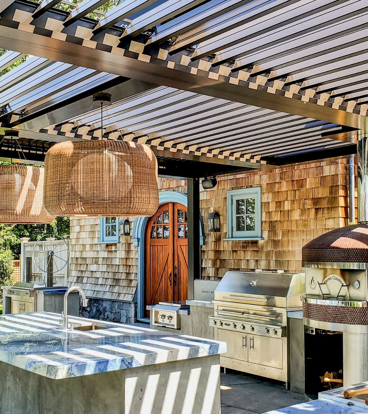 Photo of an outdoor kitchen with pizza oven, gas grill, and marble island under a slatted pergola beside a shingled building, daytime