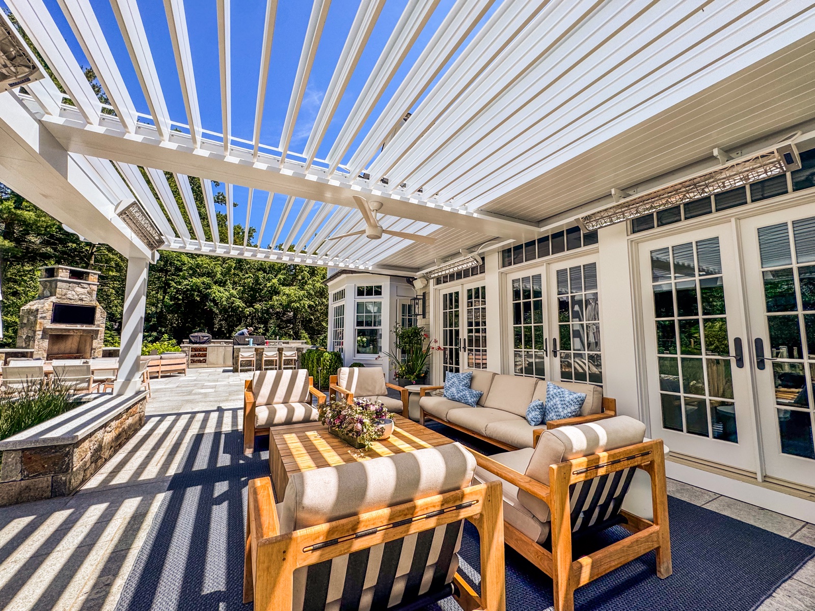 Louvered pergola lounge area with outdoor fireplace and kitchen beyond