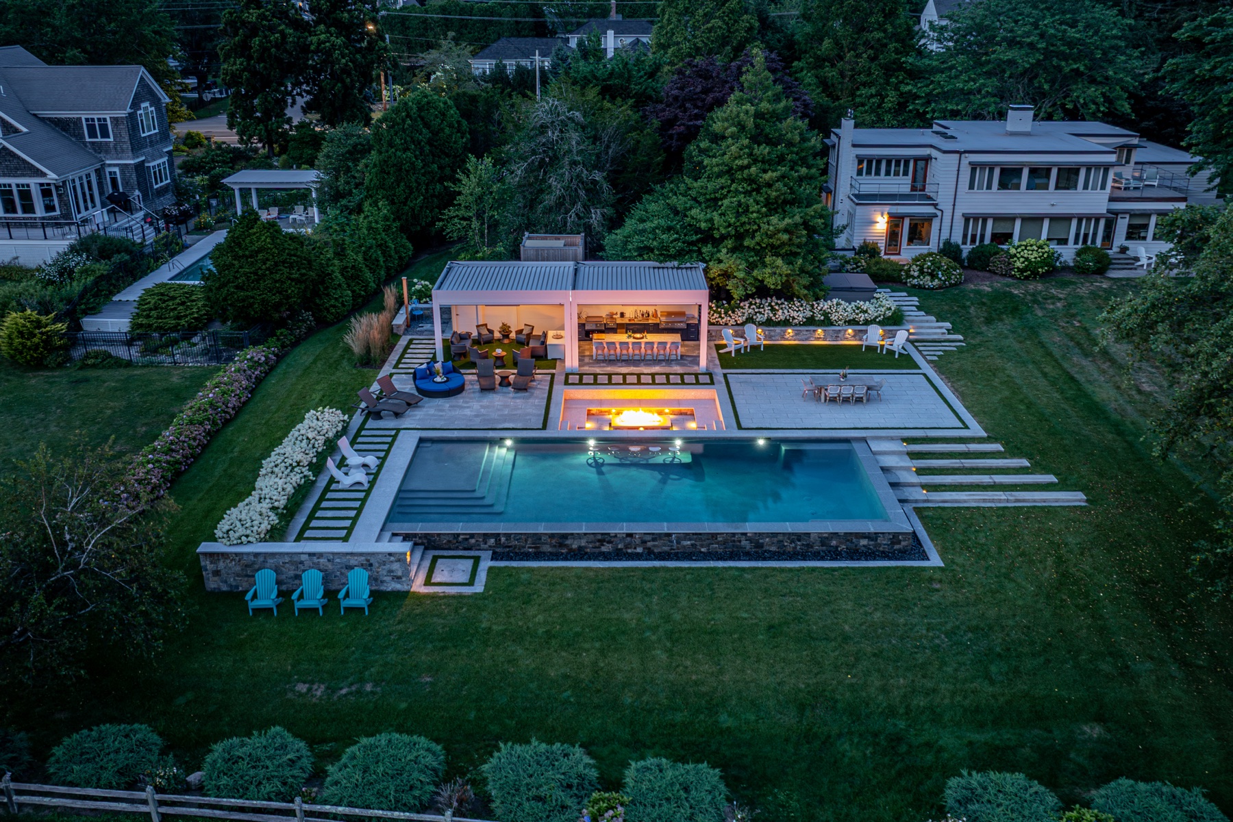 Dusk aerial of a Massachusetts estate — motorized louvered pergola, infinity-edge pool and fire feature lit at twilight