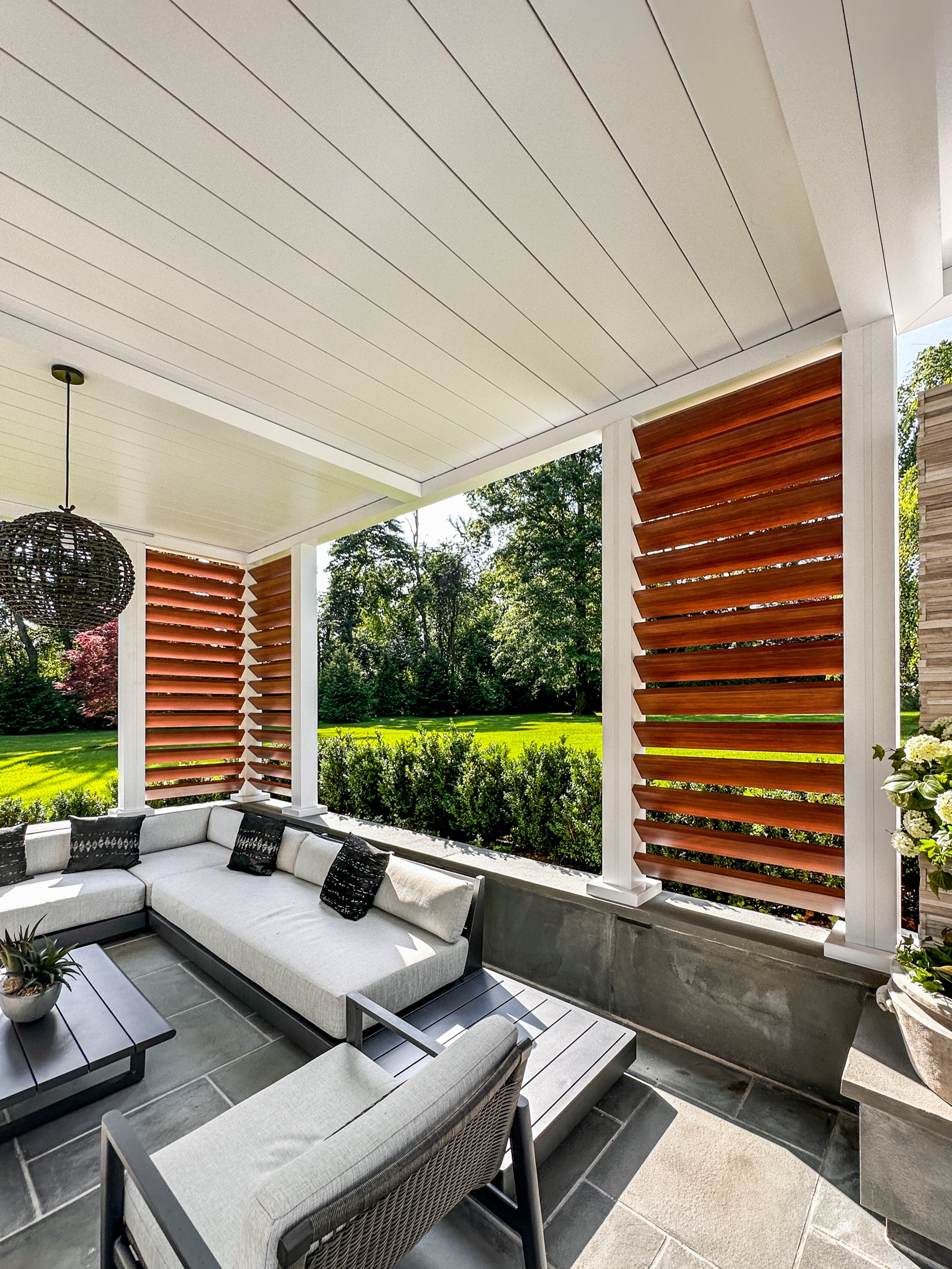 Photo of covered patio with white pergola roof, wood-tone louvered privacy screens, wicker seating, pendant lights, daytime.