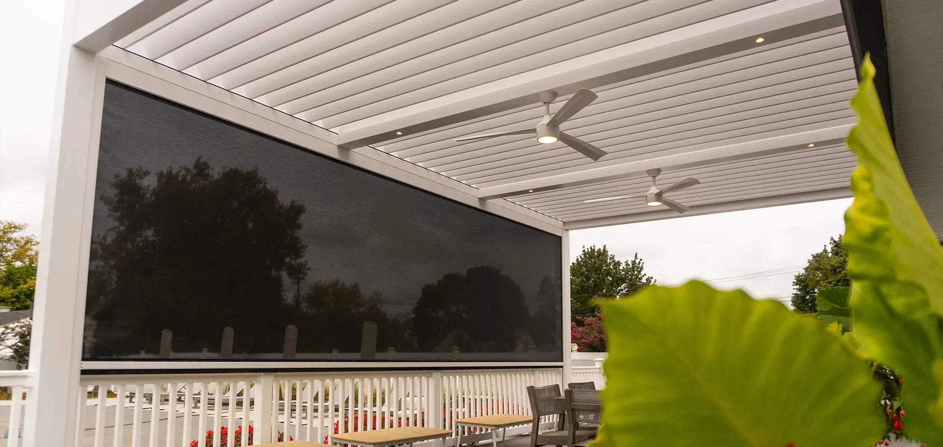 Close-up of a white louvered pergola roof with ceiling fans and a black motorized screen lowered