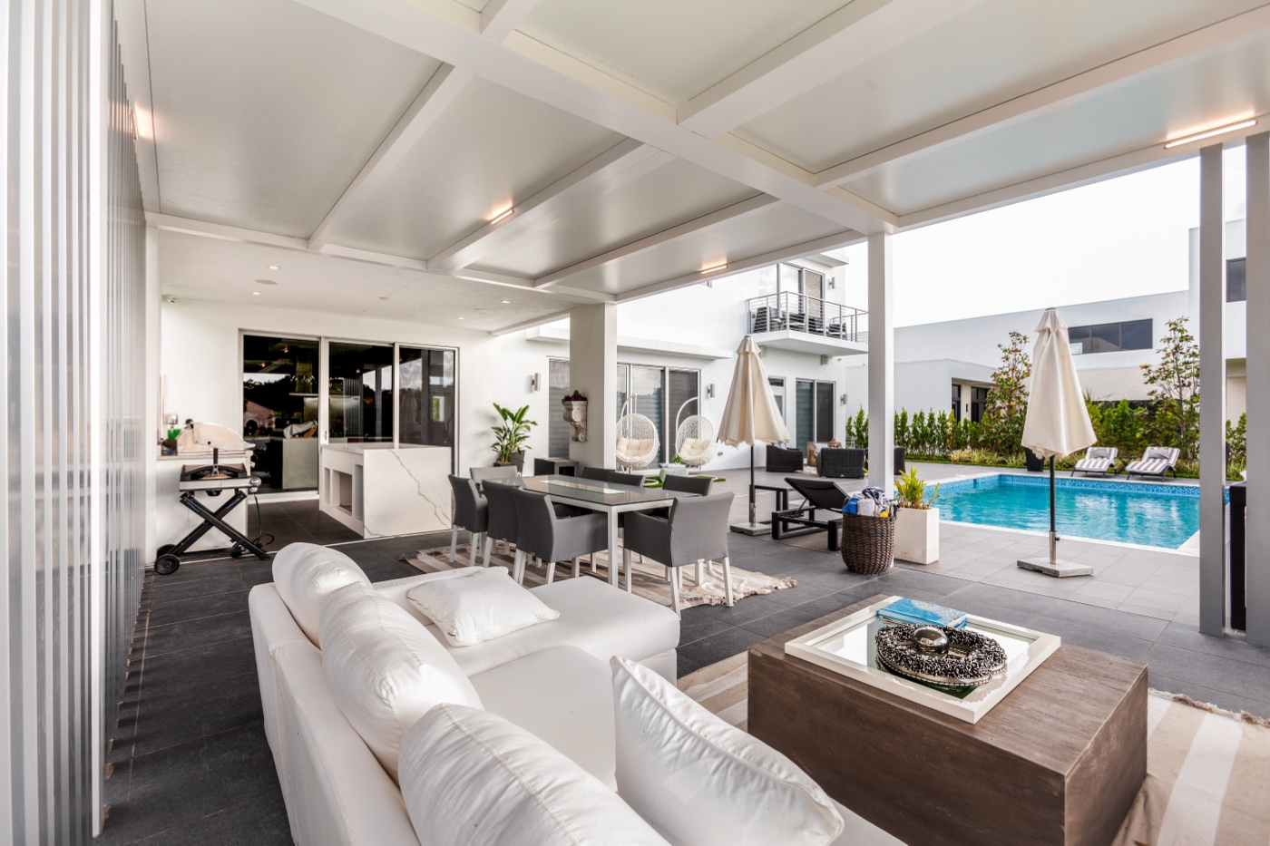 Photo of a white pergola-covered outdoor kitchen and bar with grill, ceiling fan, and stone counter in a wooded yard, daytime