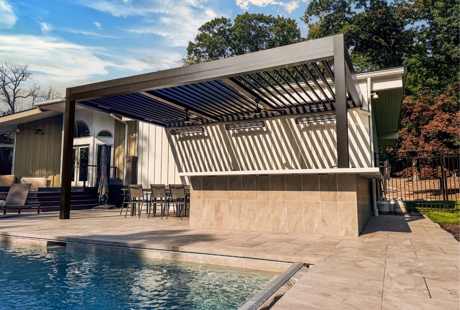 Modern patio with a retractable awning, white dining table, and chairs, bordered by greenery and a swimming pool .