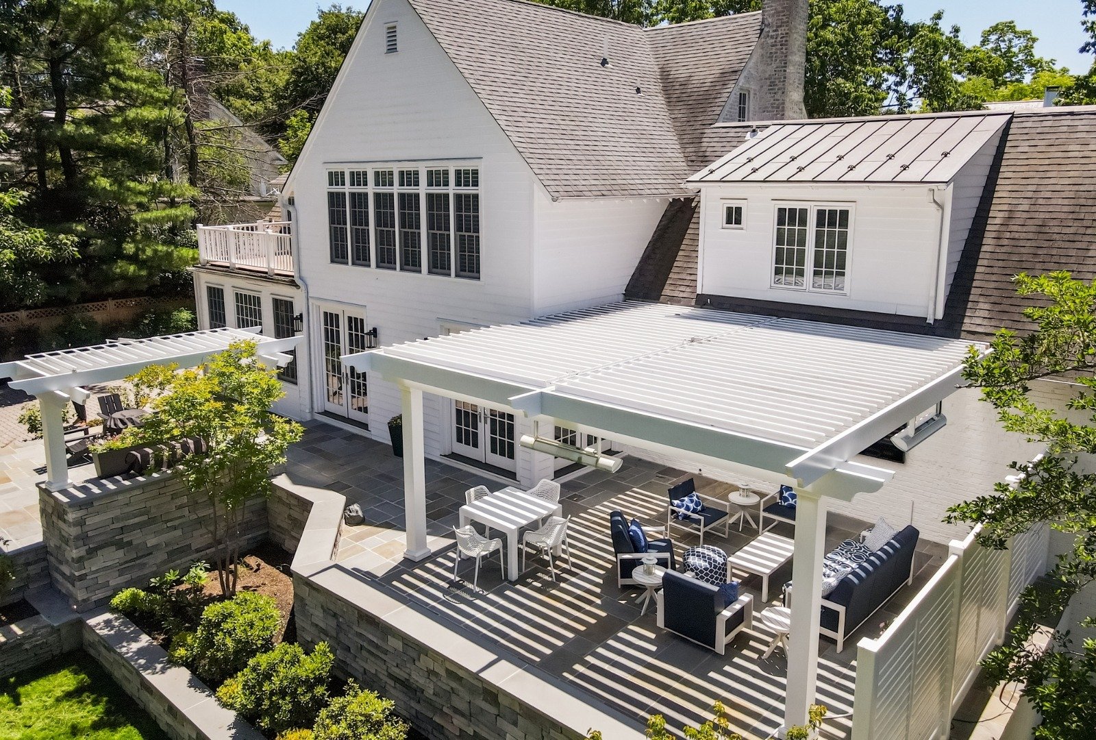 Modern patio with a retractable awning, white dining table, and chairs, bordered by greenery and a stone wall backdrop.