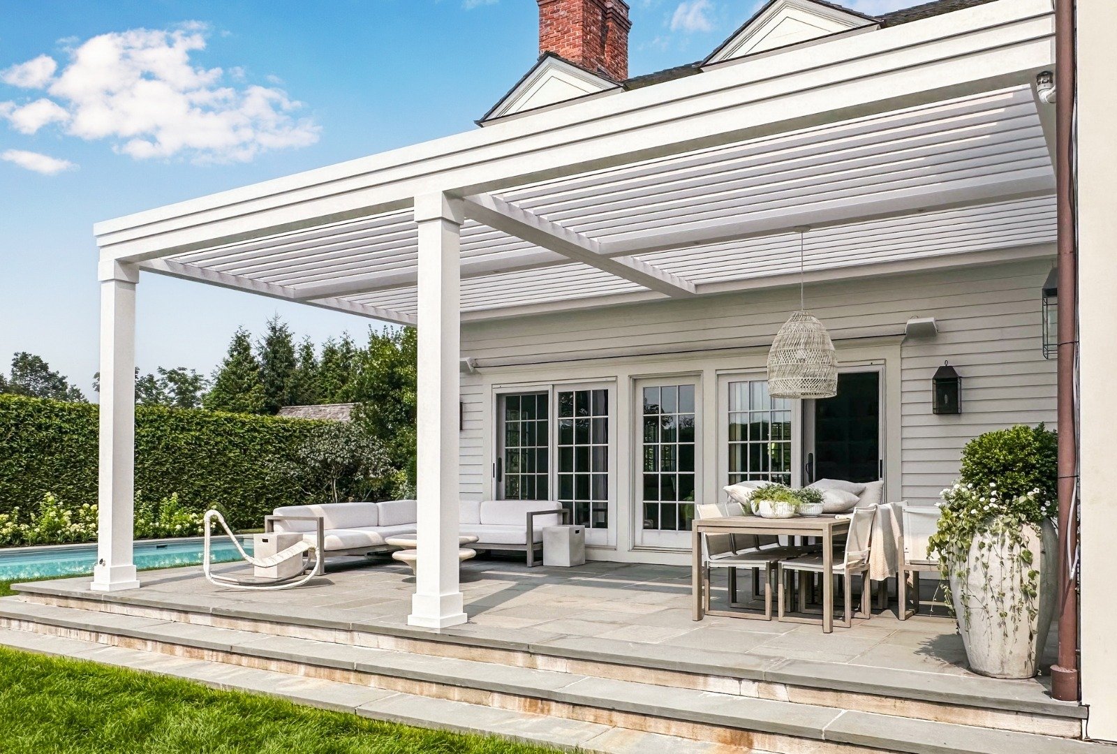 Modern patio with a retractable awning, white dining table, and chairs, bordered by greenery and a stone wall backdrop.