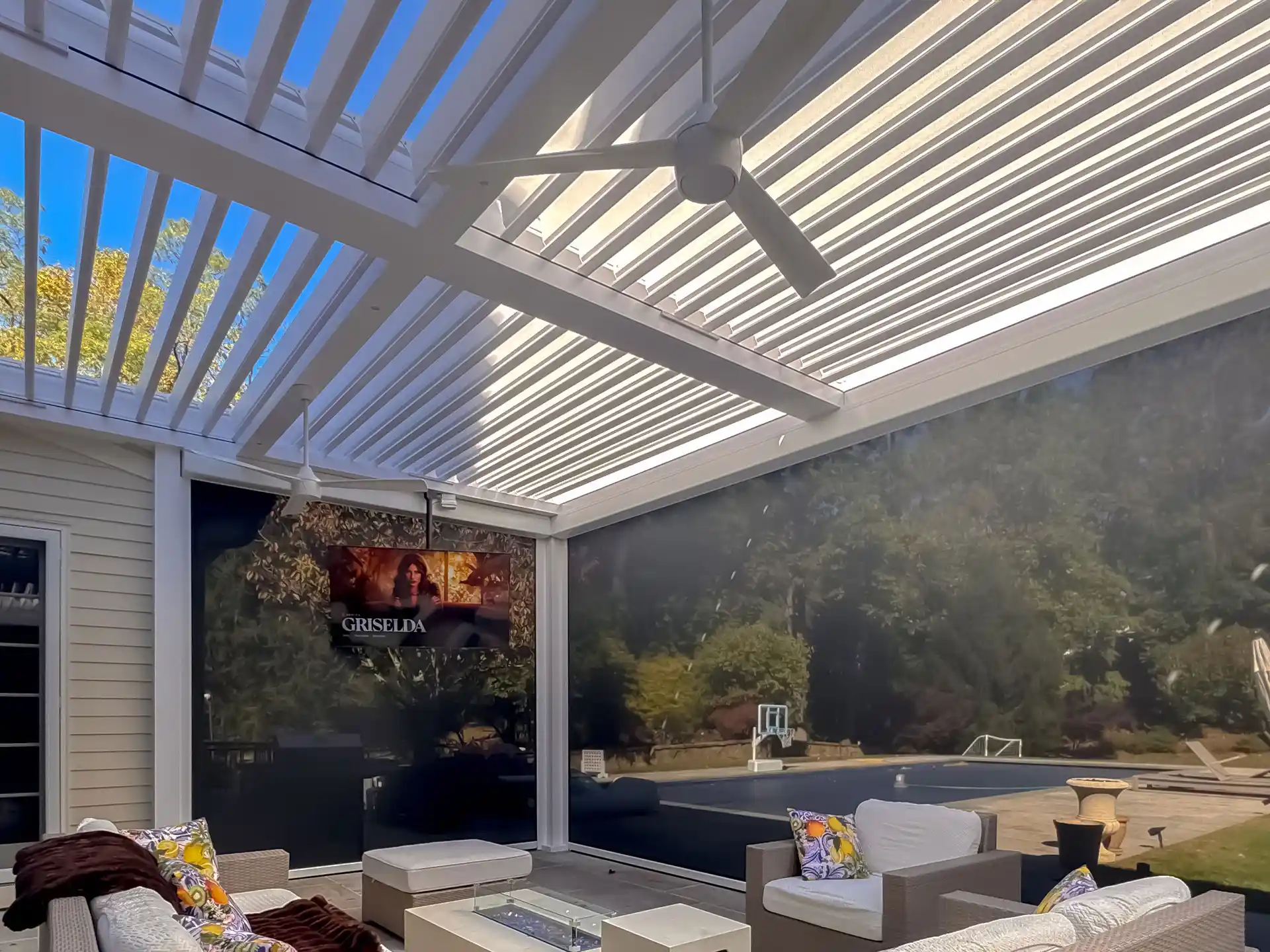 A modern outdoor patio with a pergola, ceiling fan, cozy seating, and a mounted TV, surrounded by greenery and a pool view.