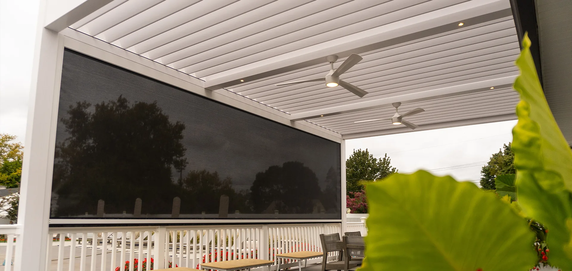 A modern, covered outdoor patio with a white pergola, ceiling fans, and a large retractable screen. The space features seating, a railing, and some greenery, creating a cozy and stylish outdoor living area 2.