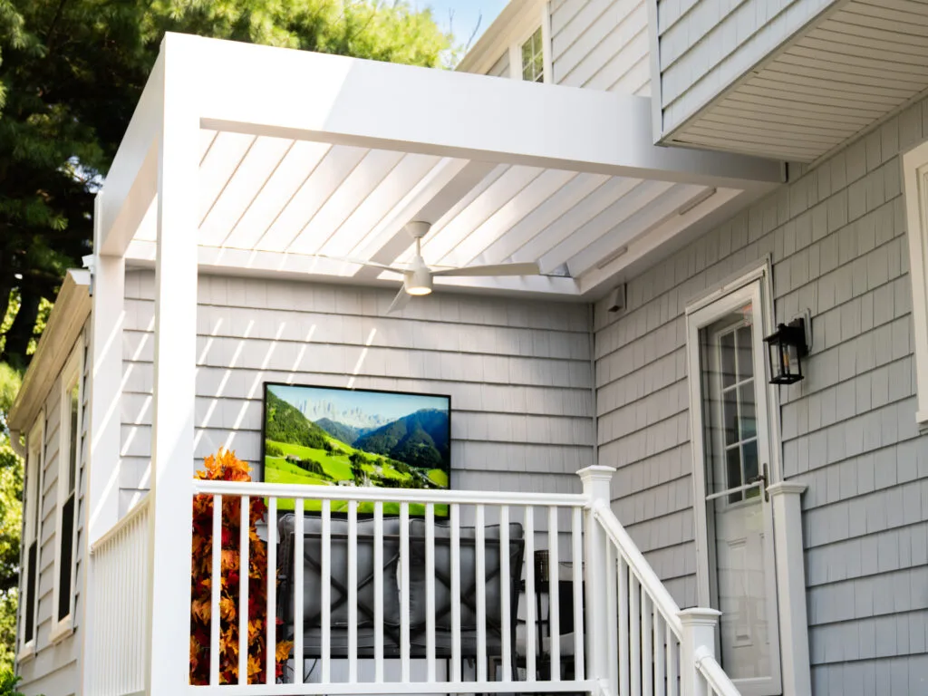 frot view of modern outdoor space with a white pergola and ceiling fan. A flat-screen TV displays nature scenery and is mounted on light gray shiplap siding. A railing with decorative foliage is in the foreground.