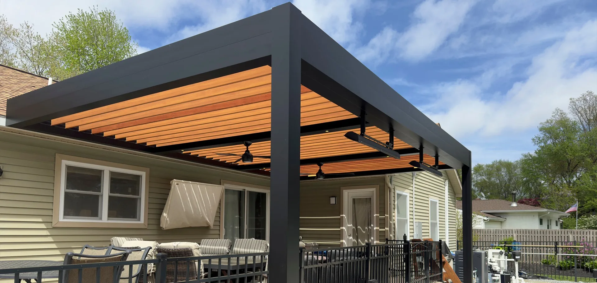 A modern patio with a black pergola featuring wooden slats, overlooking a backyard with lush greenery and a serene sky.