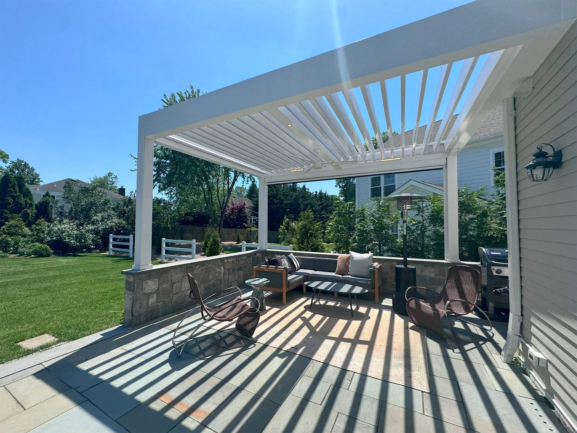 Sunny outdoor patio featuring a modern gray sectional, decorative pillows, a stone table, and a pergola above with slatted roof.