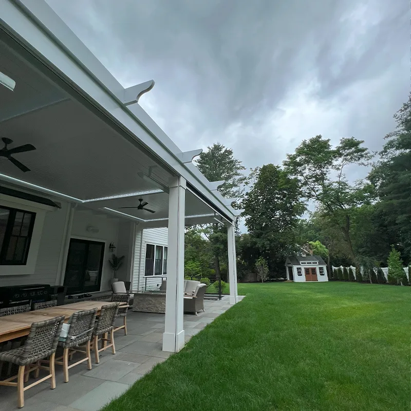 Covered patio features a wooden dining table, wicker chairs, ceiling fans, and a view of a lush green yard with a garden shed.
