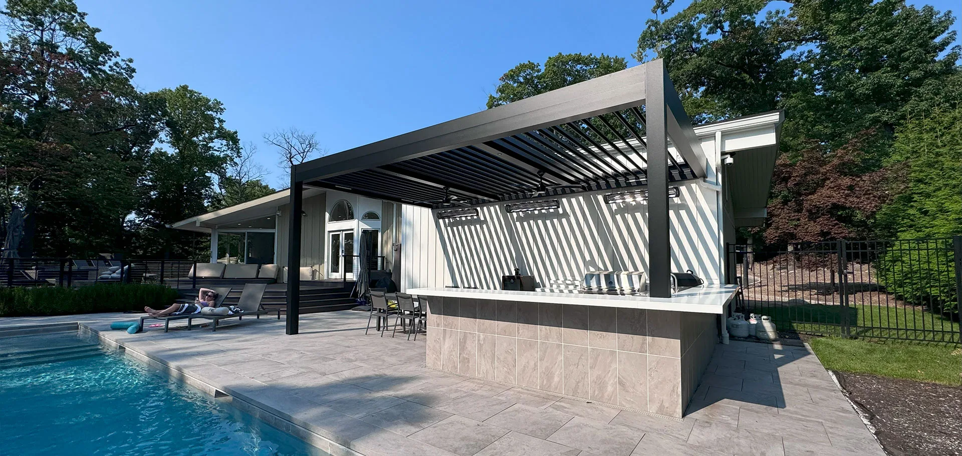 A modern backyard with a swimming pool and a covered patio area. The patio has a pergola with slatted roofing, providing shade.