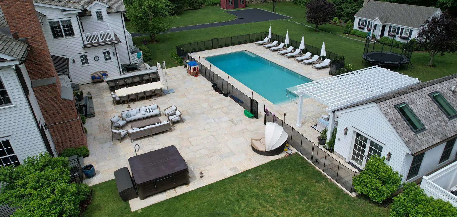Aerial view of a backyard featuring a large swimming pool surrounded by lounge chairs and white umbrellas. Theres a pergola, a trampoline, and outdoor seating areas. The scene includes a grassy lawn and nearby buildings.
