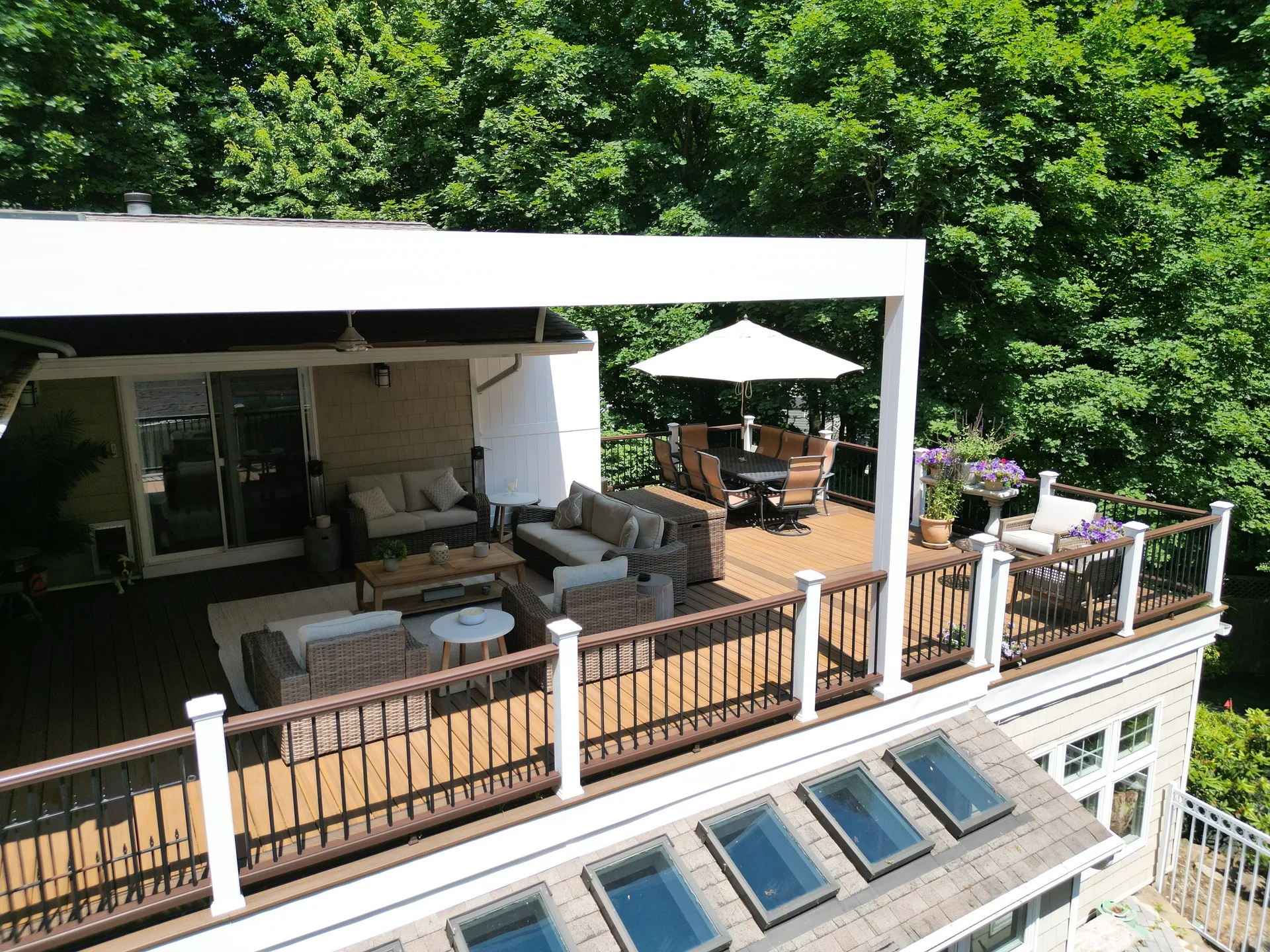 Side view of modern outdoor living space with a covered patio, comfortable furniture, and a man standing near a TV amidst lush green surroundings.