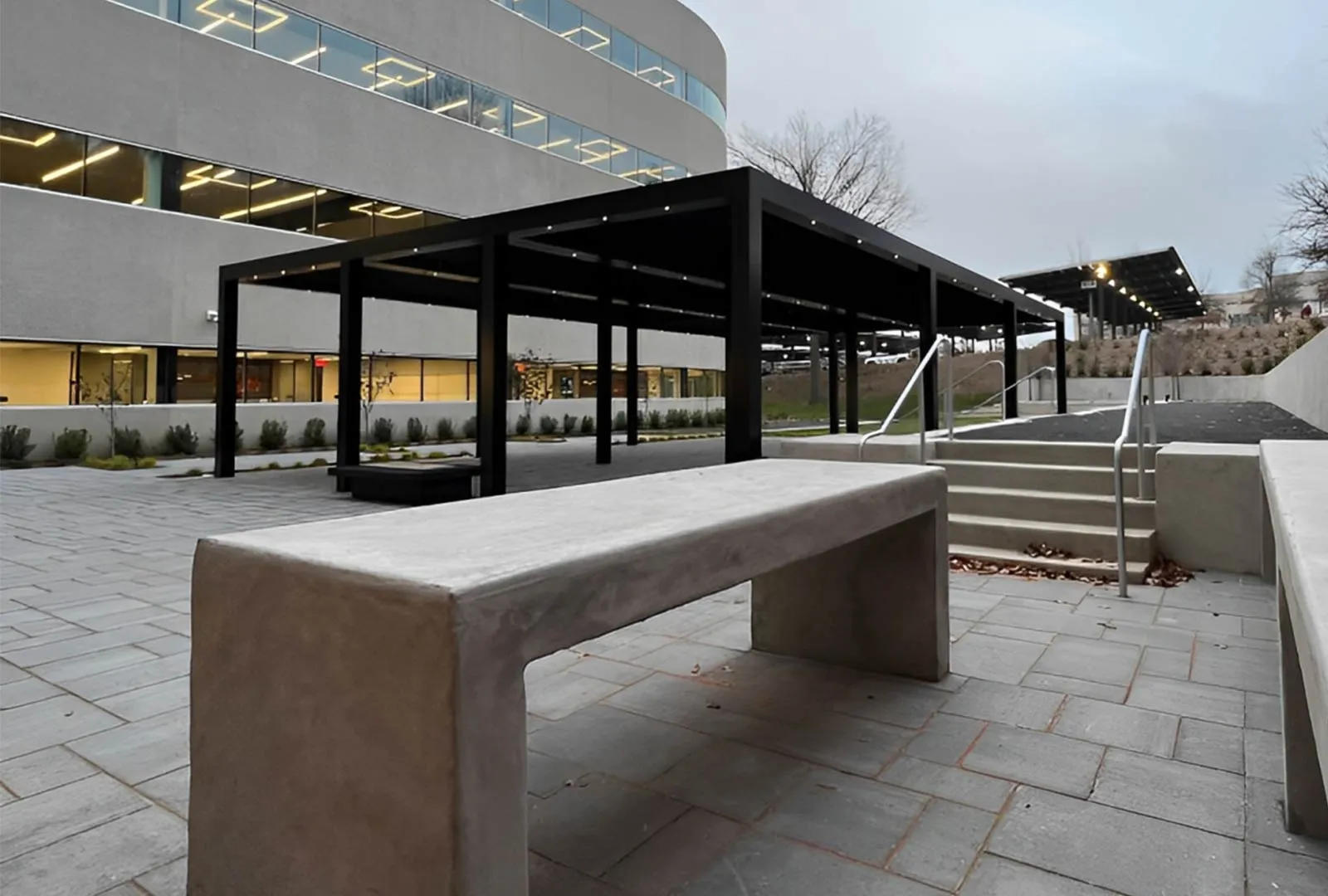A modern outdoor space featuring a concrete bench, black canopy, and steps leading to a landscaped area beside a contemporary building.
