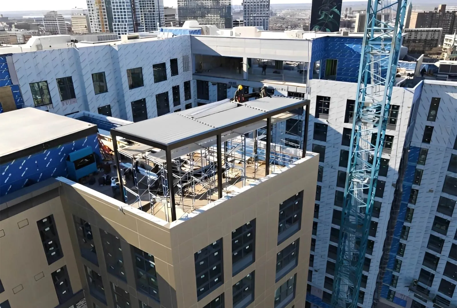 Construction workers install a steel frame on a partially completed rooftop terrace, surrounded by a high-rise urban skyline.
