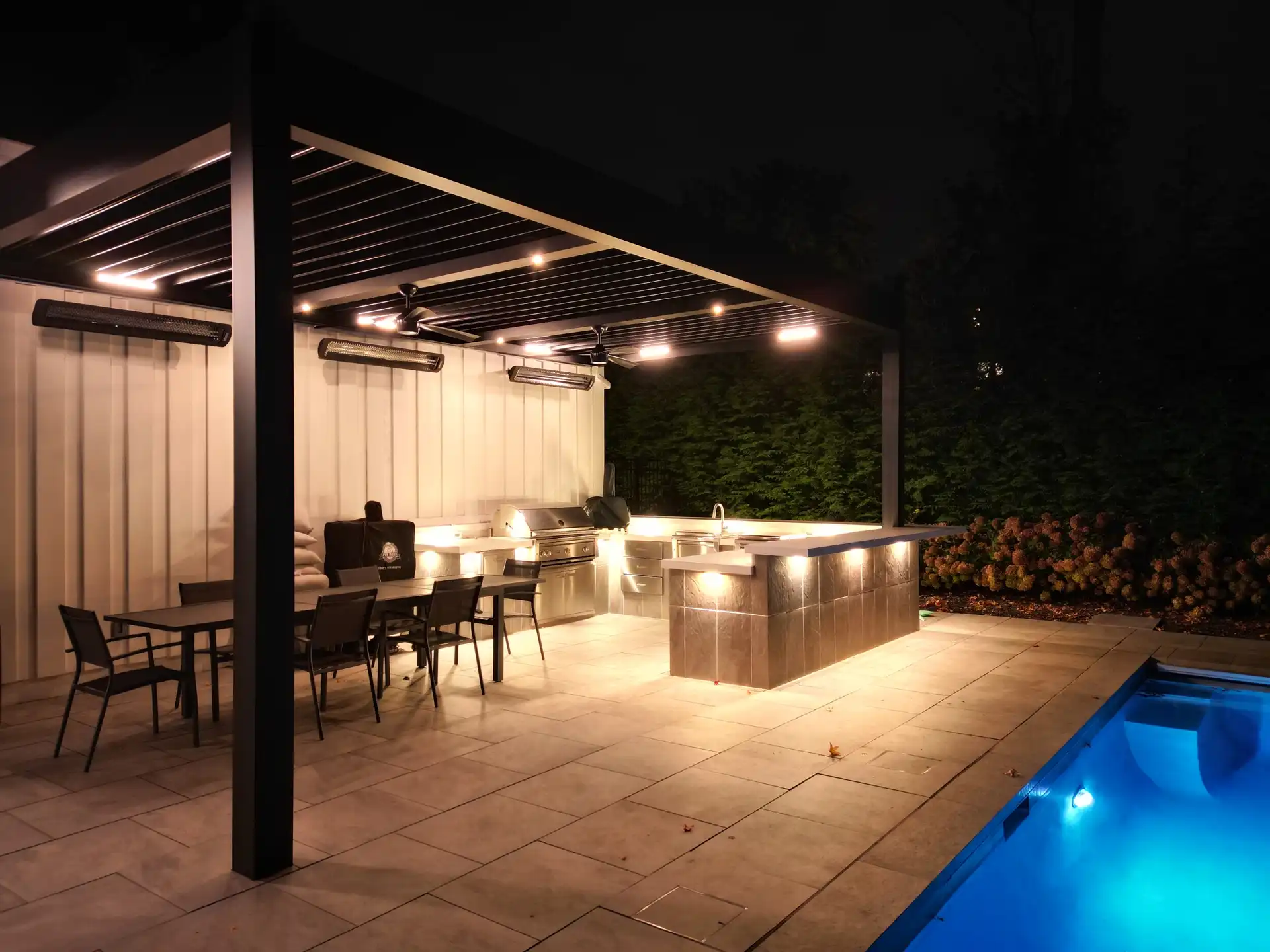 Modern outdoor kitchen at night with glowing lights, sleek counters, and a dining table under a pergola. The area features a grill and sinks, bordered by a swimming pool and lush greenery2.